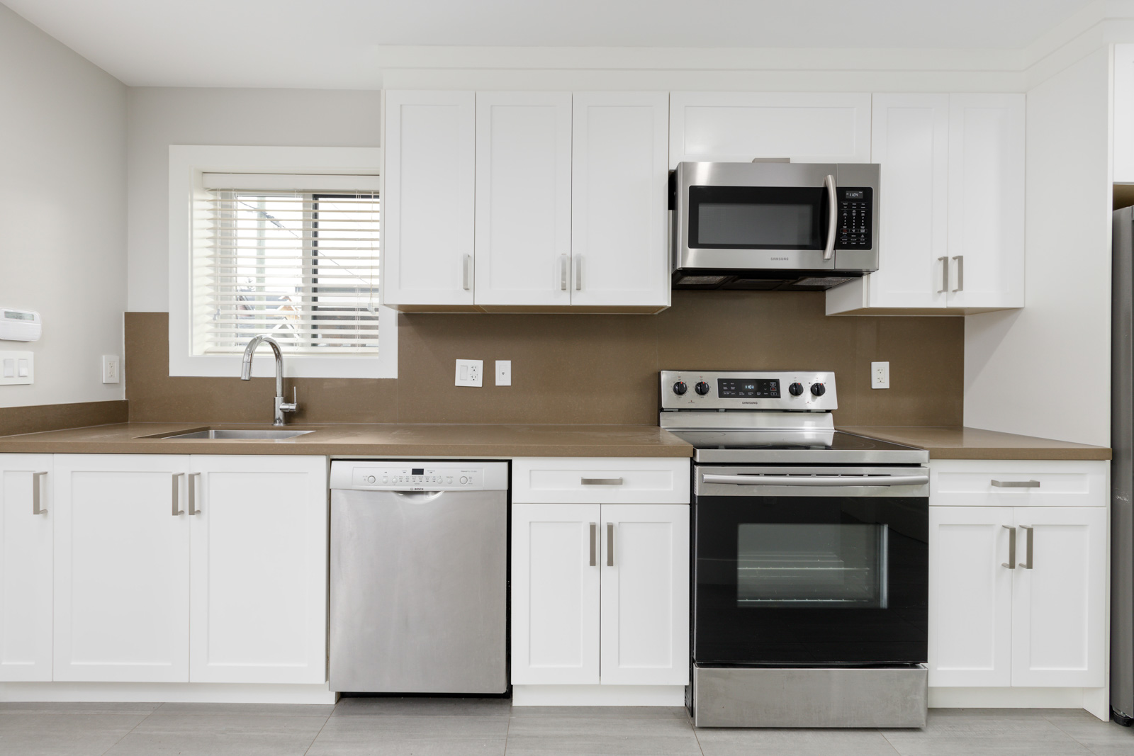 Modern kitchen with white cabinets, stainless steel appliances including a dishwasher, oven, and microwave, a brown backsplash, and a window above the sink.
